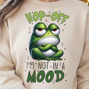 May include: Beige sweatshirt with a green frog graphic. The frog has an angry expression with arms crossed. Text above the frog reads "HOP-OFF" and below reads "I'M NOT IN A MOOD". Small stars surround the text and frog.