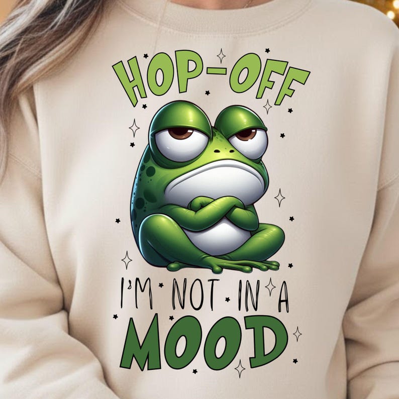 May include: Beige sweatshirt with a green frog graphic. The frog has an angry expression with arms crossed. Text above the frog reads "HOP-OFF" and below reads "I'M NOT IN A MOOD". Small stars surround the text and frog.