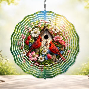 May include: A decorative wind spinner featuring a birdhouse scene with two red cardinals, surrounded by colorful flowers. The spinner has a wavy, green outer edge and is designed to hang outdoors.