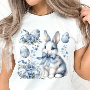 May include: White t-shirt featuring a blue bunny rabbit wearing a blue bow and surrounded by blue and white Easter eggs and flowers.