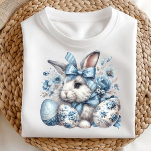 May include: White t-shirt with a blue and white Easter bunny design. The bunny is wearing a blue bow and is surrounded by blue and white flowers and eggs.