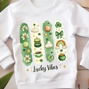 Puede incluir: Sudadera blanca con un diseño verde, dorado y blanco que presenta un arcoíris, tréboles, nubes, tazas de café, macarons y una olla de oro. El texto "Lucky Vibes" está impreso en dorado en la sudadera.
