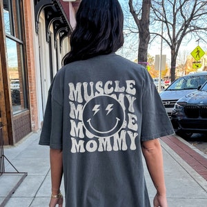 K&ouml;nnte beinhalten: &Uuml;berdimensionales T-Shirt in Pfefferfarbe mit dem Text "MUSCLE MOMMY" in einem Retro-Schriftzug, der ein l&auml;chelndes Gesicht mit Blitzaugen umgibt. Das Shirt ist ein l&auml;ssiger, bequemer Stil.