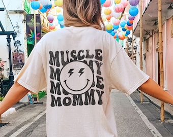 Może przedstawiać: Kremowa koszulka z napisem "MUSCLE MOMMY" i uśmiechniętą buzią z błyskawicą. Koszulka ma swobodny styl, idealna do noszenia na co dzień.