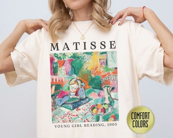 May include: A white t-shirt featuring a colourful painting of a young girl reading by Henri Matisse. The painting is titled "Young Girl Reading, 1905". The text "Matisse" is printed above the painting. The t-shirt has a "Comfort Colors" label.