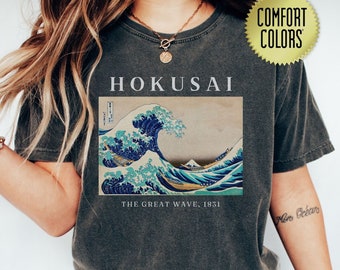 May include: Dark gray t-shirt featuring a print of Hokusai's "The Great Wave off Kanagawa" artwork. The design includes the text "HOKUSAI" above the image and "THE GREAT WAVE, 1831" below. The shirt has a "Comfort Colors" tag.