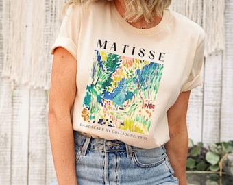 May include: A soft cream colored t-shirt with a colorful painting of a landscape by Henri Matisse. The text "MATISSE" and "LANDSCAPE AT COLLIOURE, 1905" is printed on the shirt.