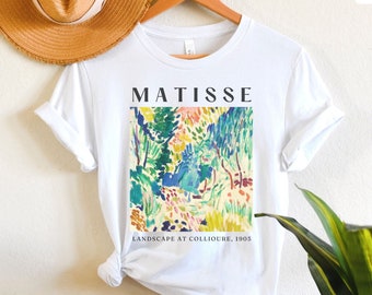 Może przedstawiać: Biała koszulka z kolorowym nadrukiem pejzażu autorstwa Matisse'a. Nadruk zawiera słowo "MATISSE" nad grafiką i "LANDSCAPE AT COLLIOURE, 1905" poniżej. Koszulka wisi na drewnianym wieszaku.