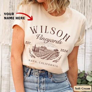 May include: A soft cream colored t-shirt with a brown graphic design that says "Wilson Vineyards Est. 2024 Napa, California".