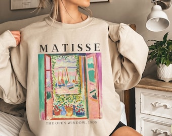 Może przedstawiać: Beżowa bluza z nadrukiem graficznym obrazu Henri Matisse'a zatytułowanego "Otwarte okno, 1905". Obraz przedstawia okno z widokiem na port i łodzie. Bluza ma okrągły dekolt i długie rękawy.