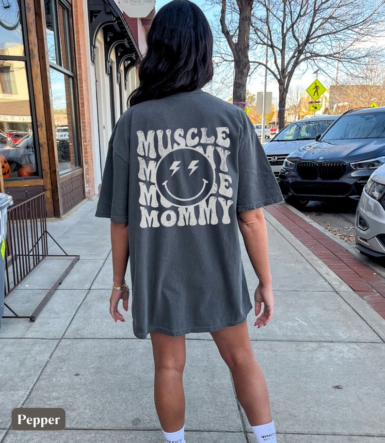 K&ouml;nnte beinhalten: &Uuml;berdimensionales T-Shirt in Pfefferfarbe mit dem Text "MUSCLE MOMMY" in einem Retro-Schriftzug, der ein l&auml;chelndes Gesicht mit Blitzaugen umgibt. Das Shirt ist ein l&auml;ssiger, bequemer Stil.