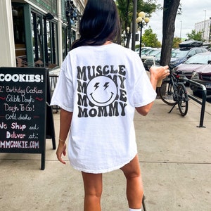 K&ouml;nnte beinhalten: Wei&szlig;es T-Shirt mit dem Text "MUSCLE MOMMY" in Retro-Schrift, umgeben von einem l&auml;chelnden Gesicht mit Blitzaugen. Die Person steht drau&szlig;en in der N&auml;he eines Schildes mit Werbung f&uuml;r Kekse.