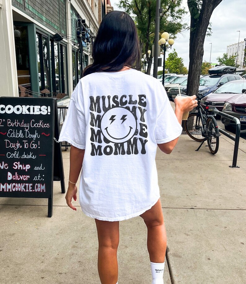 K&ouml;nnte beinhalten: Wei&szlig;es T-Shirt mit dem Text "MUSCLE MOMMY" in Retro-Schrift, umgeben von einem l&auml;chelnden Gesicht mit Blitzaugen. Die Person steht drau&szlig;en in der N&auml;he eines Schildes mit Werbung f&uuml;r Kekse.