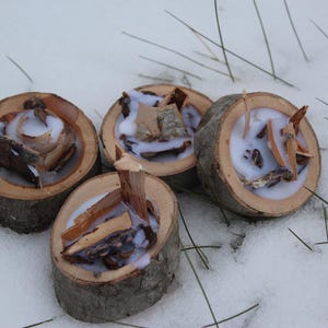 May include: Four rustic candles made from wood slices, each filled with white wax and pieces of bark. The candles are arranged on a snowy surface, creating a natural and cozy aesthetic. The wood slices are approximately 2-3 inches in diameter.