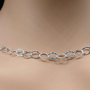 May include: A silver necklace featuring a hammered chain design. The chain is made up of oval links, giving a textured and reflective appearance. The necklace is shown against a neutral backdrop, emphasising its simple elegance.