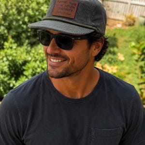 Puede incluir: Una gorra de béisbol de pana gris carbón con un parche rectangular de cuero marrón en la parte delantera. El parche tiene grabado el texto SUPPORT YOUR LOCAL AUTISM COMMUNITY y Aloha Autism. La gorra incluye un detalle de cordón gris.
