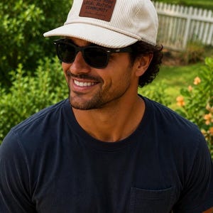 Puede incluir: Una gorra de béisbol de pana color crema con un parche rectangular de cuero marrón en la parte delantera. El parche tiene grabado el texto SUPPORT YOUR LOCAL AUTISM COMMUNITY y Aloha Autism. La gorra presenta una corona estructurada y visera.