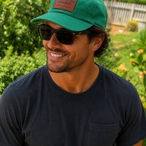 Puede incluir: Una gorra de béisbol verde con un parche rectangular de cuero marrón en la parte delantera. El parche tiene grabado el texto SUPPORT YOUR LOCAL AUTISM COMMUNITY y las palabras Aloha Autism en letra cursiva debajo del mensaje principal.