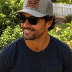 Puede incluir: Una gorra tipo camionero gris con parte trasera de malla y un parche rectangular de cuero marrón en la parte delantera. El parche presenta el texto negro "SUPPORT YOUR LOCAL AUTISM COMMUNITY" seguido del texto "Aloha Autism" en letra cursiva.