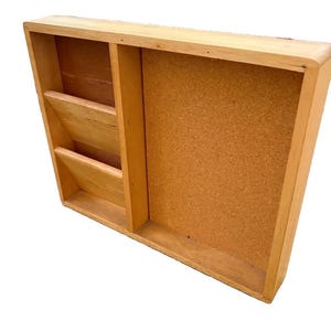May include: A rectangular wall-mounted organizer crafted from light-toned wood, featuring two stacked open storage compartments on the left side and a large, vertical rectangular cork board panel on the right side for pinning notes or documents.