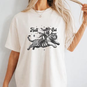 Könnte beinhalten: Cremefarbenes T-Shirt mit einer Schwarz-Weiß-Grafik einer Person, die auf einem Tiger reitet. Der Text "Hell Cat" befindet sich über dem Bild. Das Design umfasst Sterne und einen fließenden Schal, was eine dynamische und kantige Ästhetik erzeugt.