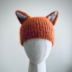 May include: A knitted fox hat with orange and grey fur. The hat is on a white mannequin head.