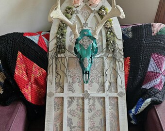 Puede incluir: Un marco de madera pintado de blanco con un diseño de arco gótico. El marco está decorado con vegetación artificial, flores rosas y una calavera de ciervo turquesa con detalles plateados. El marco está sentado en un sofá morado.