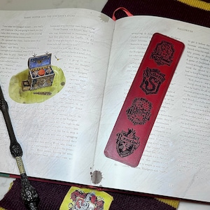 May include: A close-up of a red bookmark with black and white illustrations of the four Hogwarts houses: Gryffindor, Slytherin, Hufflepuff, and Ravenclaw. The bookmark is lying on an open book with the text "HARRY POTTER AND THE SORCERER'S STONE" visible.