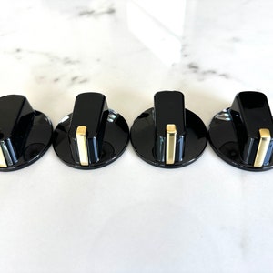 May include: Four black appliance knobs with a glossy finish and gold-colored accents. Each knob has a rounded base and a rectangular handle. The knobs are arranged in a row on a white marble surface.