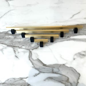 May include: A set of five gold cabinet pulls with black rectangular bases. The pulls are arranged in a pyramid shape, with the largest pull at the top and the smallest pull at the bottom.