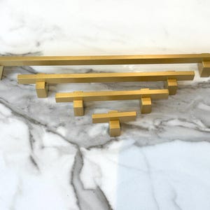 May include: A set of five gold-toned cabinet pulls in various sizes. The pulls are rectangular with square ends.