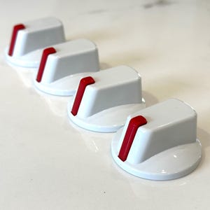 May include: Four white stove knobs with red accents. The knobs are round with a raised, rectangular top.
