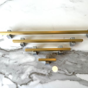 May include: Four gold and silver cabinet pulls of different sizes. The pulls are rectangular with a square end cap. The pulls are arranged in order of size, with the largest pull at the top and the smallest pull at the bottom.