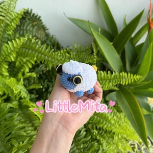 May include: A handmade crochet sheep with a white fluffy body, blue face and feet, and large black and yellow eyes. The sheep is held in a hand against a backdrop of green foliage. The text "LittleMita" is visible.