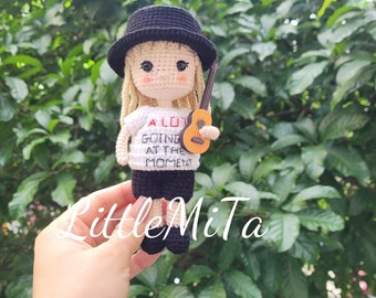 May include: A crocheted doll wearing a black hat and a white shirt with the text "A LOT GOING AT THE MOMENT". The doll is holding a small brown ukulele.