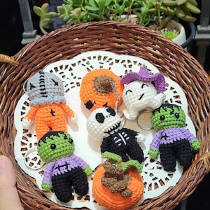 May include: A collection of seven crocheted Halloween-themed ornaments in a wicker basket. The ornaments include a pumpkin, a ghost, a skeleton, a Frankenstein, a mummy, and two green monsters.