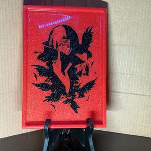 May include: A rectangular red tray with a glittery finish, featuring a black graphic of a character with crows. The tray is supported by a black stand. The design is likely inspired by anime or manga.
