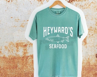 May include: A seafoam green short-sleeve t-shirt hanging on a wooden hanger. The shirt features white text that reads HEYWARD'S SEAFOOD with a line drawing of a fish centred between the two words. The fabric has a slightly faded, vintage appearance.