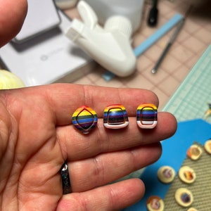 May include: Three small, colorful, rainbow-striped, resin buttons with a raised, geometric design. The buttons are in a diamond, square, and oval shape.