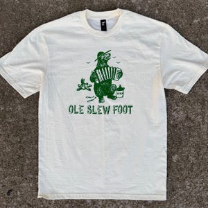 May include: Cream-colored t-shirt with a green graphic of a bear playing an accordion, wearing a hat, and holding a bottle. The text "OLE SLEW FOOT" is printed below the bear. The shirt has short sleeves and a crew neck.