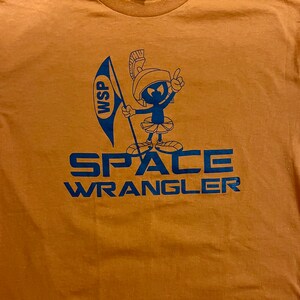 May include: A brown t-shirt with a blue cartoon character holding a flag that says "WSP". The text "SPACE WRANGLER" is printed below the character.