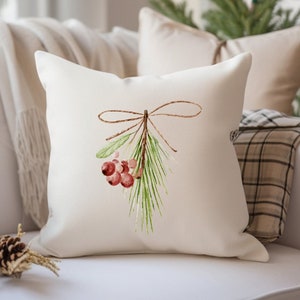 Puede incluir: Una almohada blanca con una ilustraci&oacute;n de acuarela de una rama de pino con bayas rojas atadas con una cuerda. La almohada est&aacute; en un sof&aacute; blanco con una manta de cuadros.