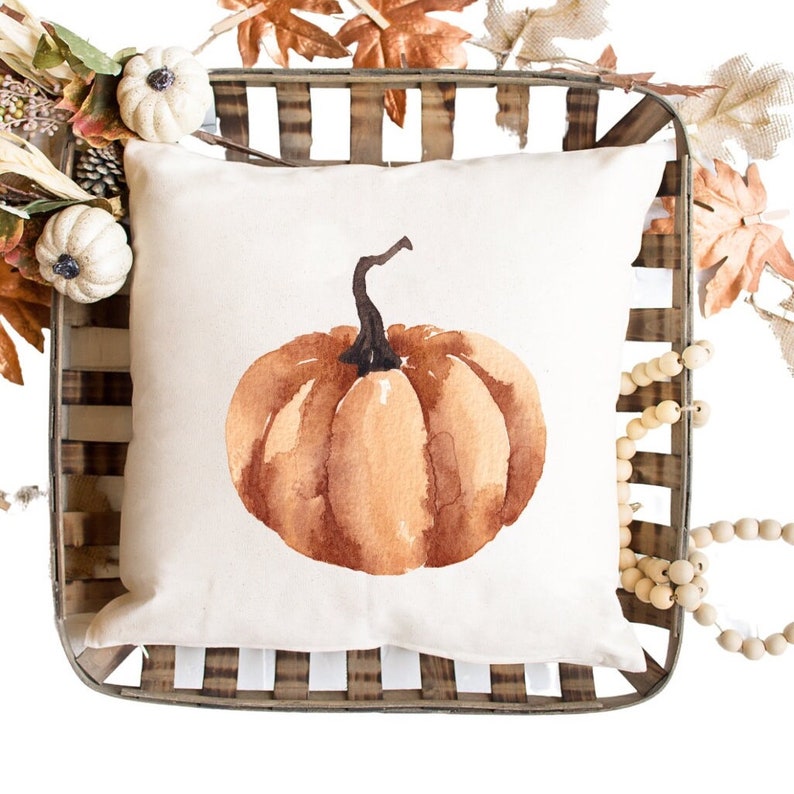 May include: A white pillow with a watercolor painting of a brown pumpkin. The pillow is in a wooden crate with fall leaves and wooden beads.