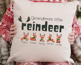 Puede incluir: Una almohada blanca con el texto "Grandma's little reindeer" en letras verdes y negras. Cinco renos de dibujos animados con gorros rojos están alineados debajo del texto, cada uno con un nombre debajo: Chris, Caleb, Kelsy, Sarah y Jody.