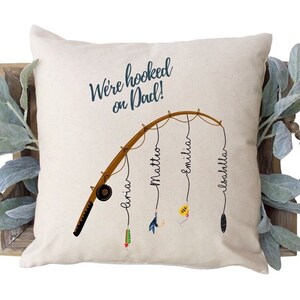 May include: A beige pillow with the text "We're hooked on Dad!" and a cartoon fishing rod with four names on it: "Amira", "Matteo", "Emilia", and "Isabella".