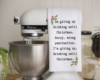 以下が含まれることがあります： 「I'm giving up drinking until Christmas. Sorry, wrong punctuation. I'm giving up. Drinking until Christmas.」のテキストが入った白いキッチンタオル。タオルにはヒイラギの実のアクセントがあり、白いスタンドミキサーにかかっています。