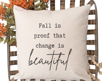May include: A white throw pillow with the text "Fall is proof that change is beautiful" printed in black. The pillow is sitting on a wooden crate with a string of wooden beads.