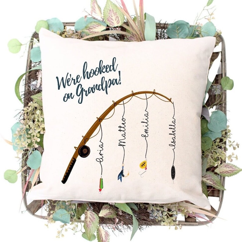 May include: A white pillow with the text "We're hooked on Grandpa!" and a cartoon fishing rod with four lures and the names "Aria", "Mattio", "Emilia", and "Isabella" written on the lines.