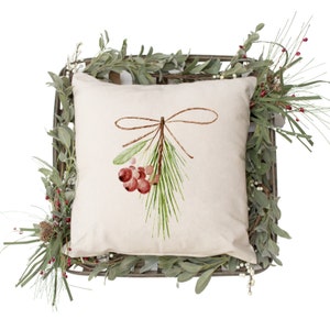 Puede incluir: Una almohada blanca con un dise&ntilde;o de acuarela de una rama de pino con bayas rojas y un lazo de cuerda marr&oacute;n.