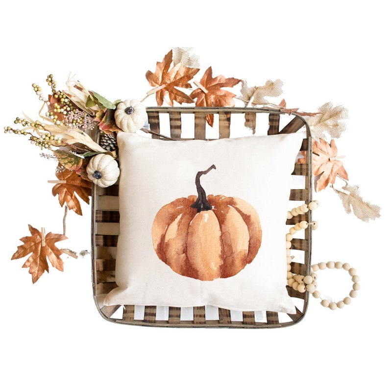 May include: A white pillow with a watercolor painting of an orange pumpkin on it. The pillow is in a wooden crate with fall leaves and white pumpkins around it.